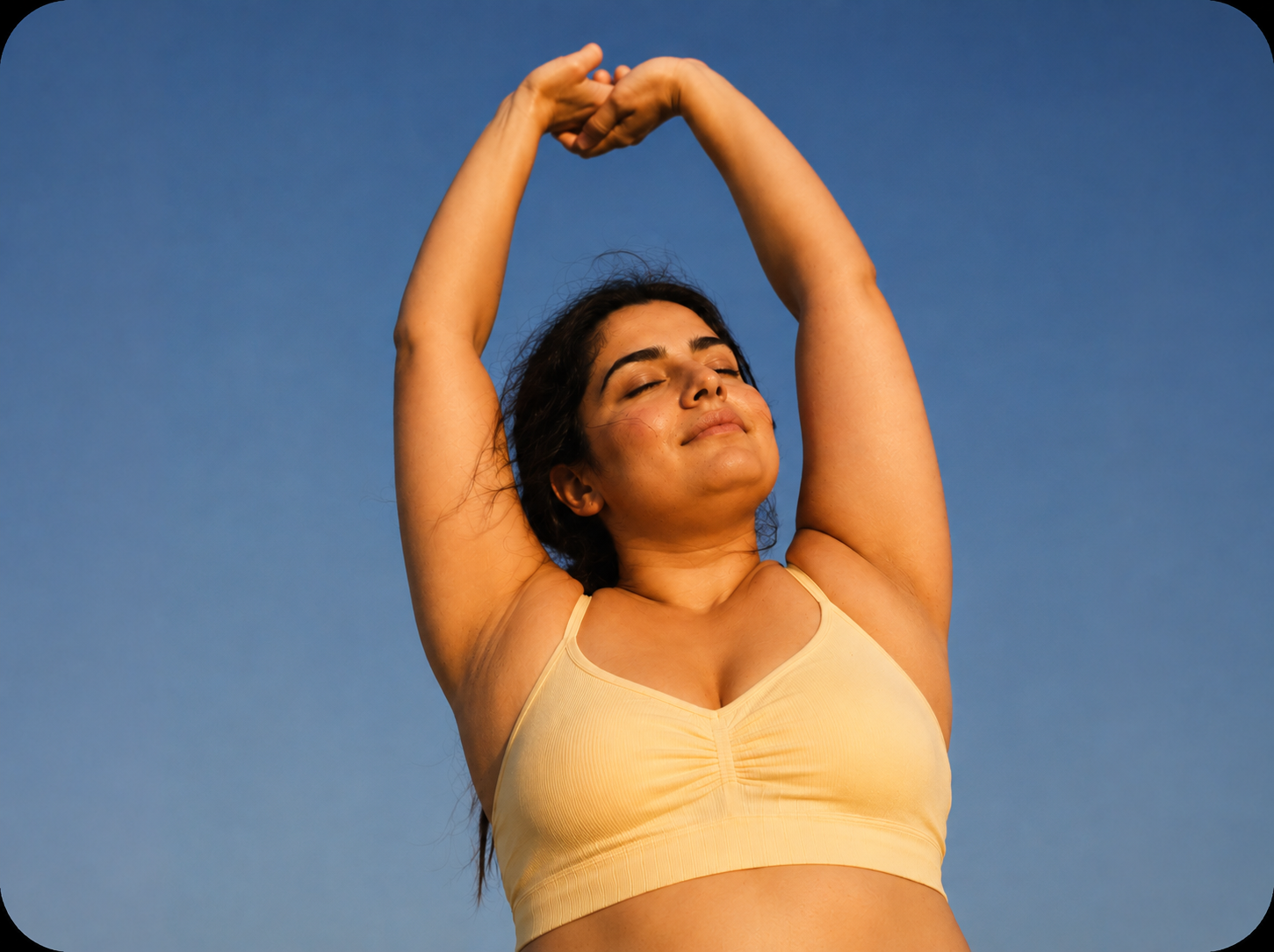 A woman stretching her arms overhead against a clear blue sky at golden hour